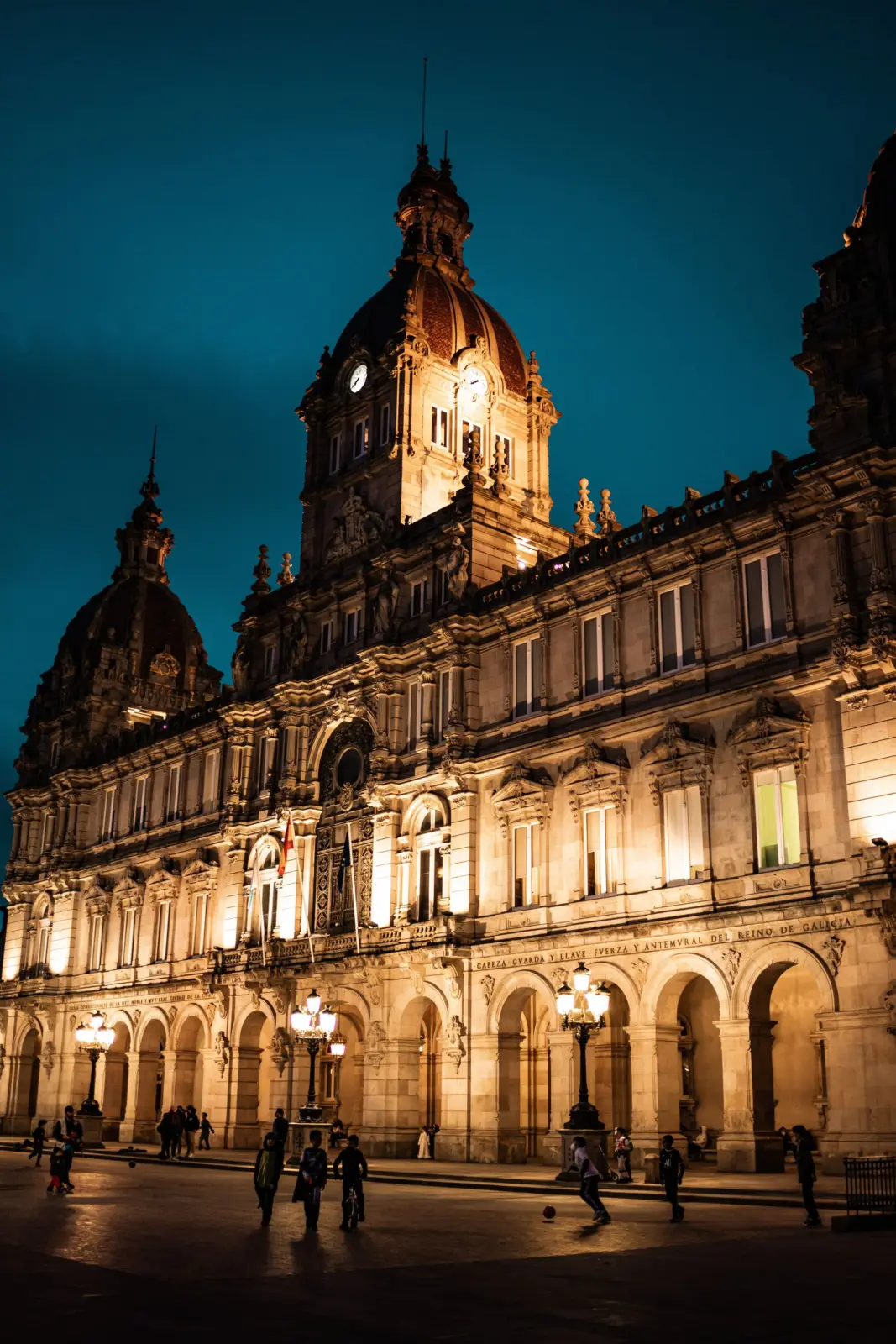 Palacio Municipal de A Coruña iluminado de noche con peatones en la plaza