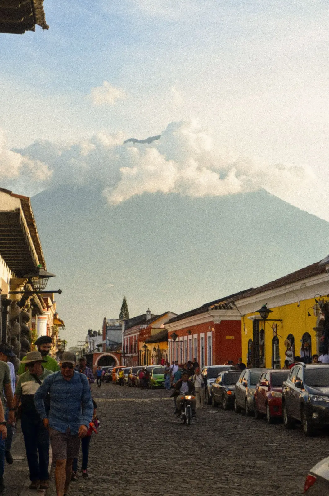 Pedestrians and vehicles on a cobblestone street in Antigua Guatemala beneath Agua Volcano