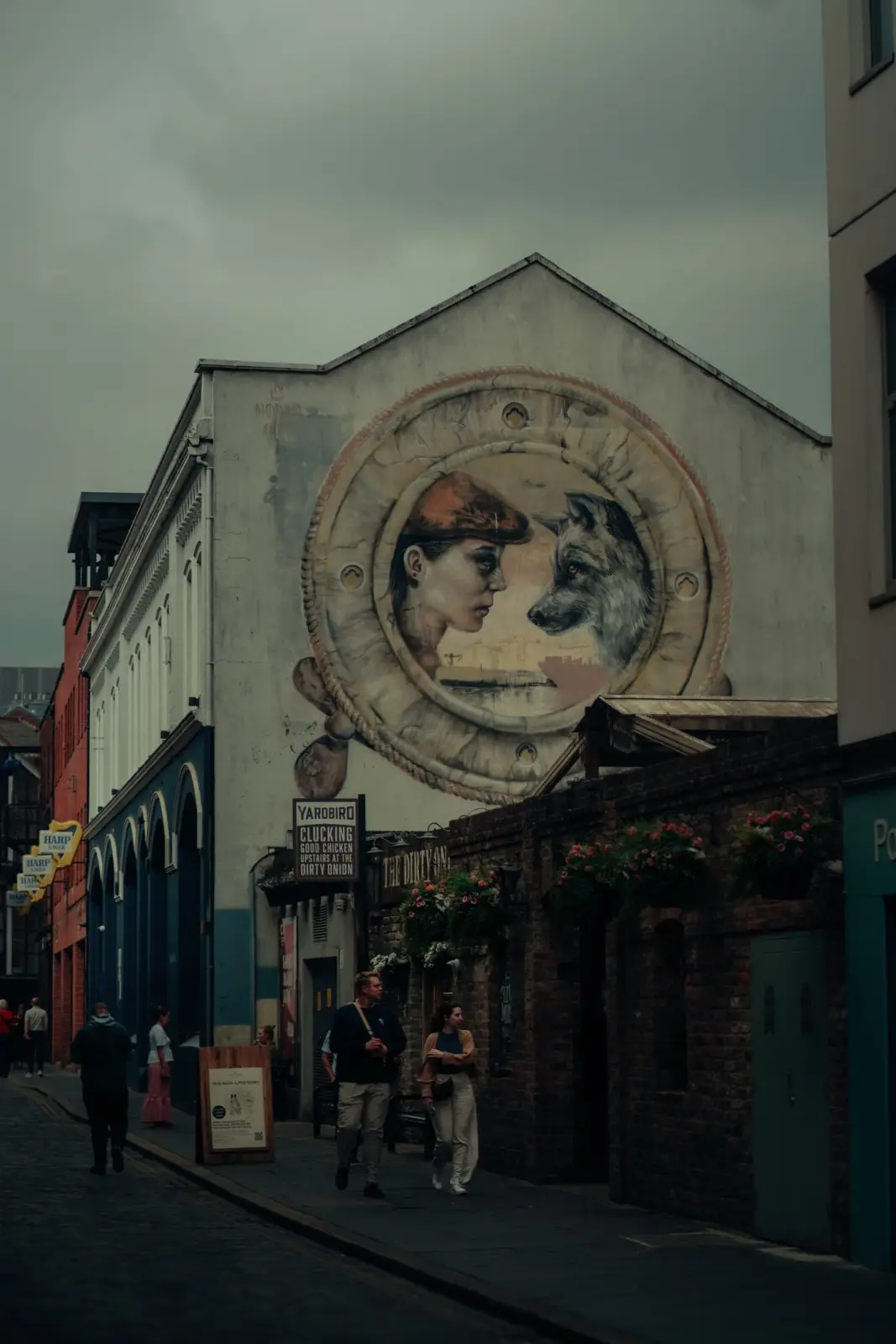 Pedestrians passing a woman-and-wolf mural in a Belfast alleyway